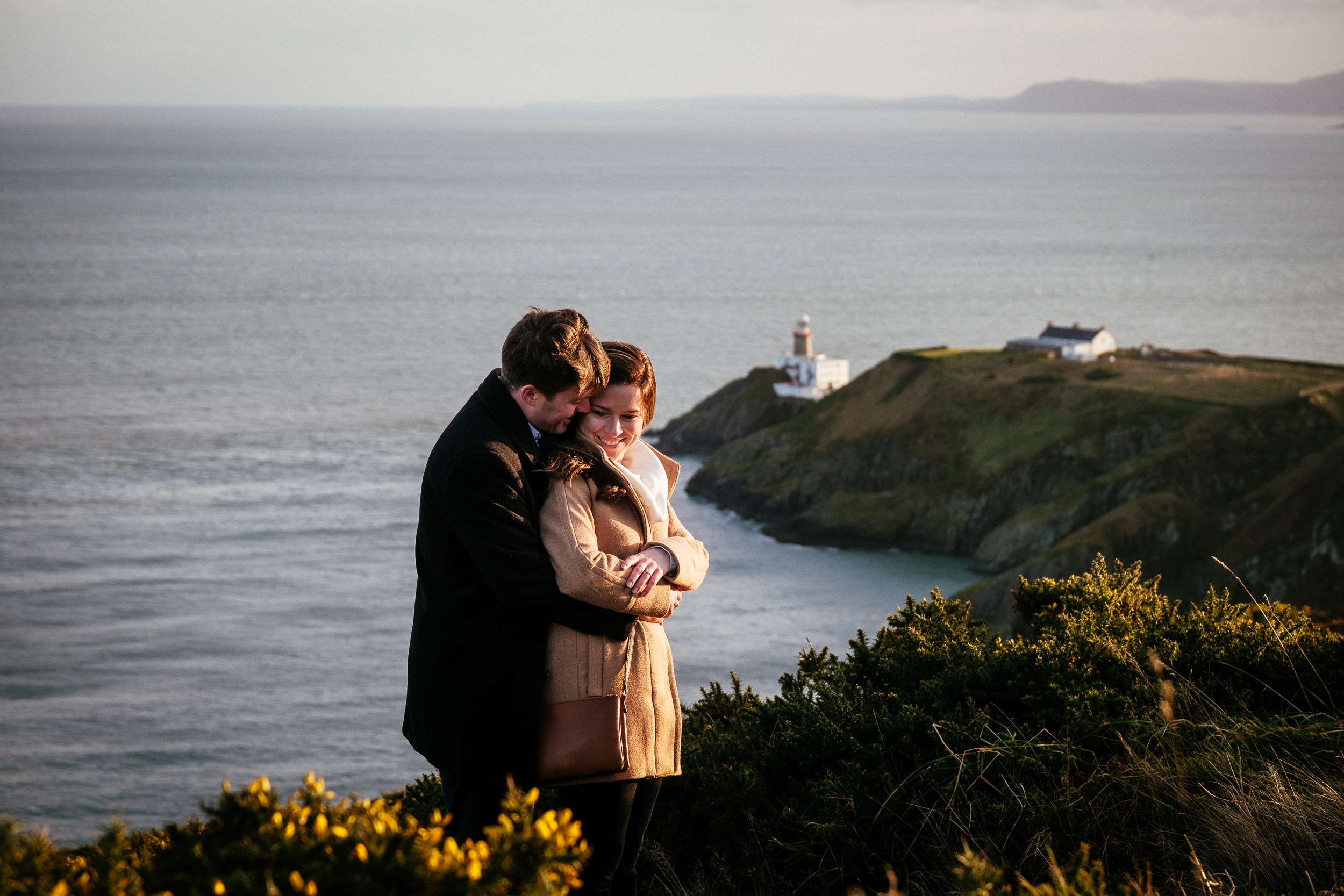 engaged couple hugging on the cliff path at Howth Summit in Dublin with teh Bailey lighthouse in the background